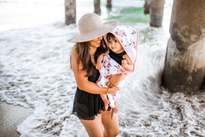 Mum wearing hat and togs at the beach with baby in a hooded towel